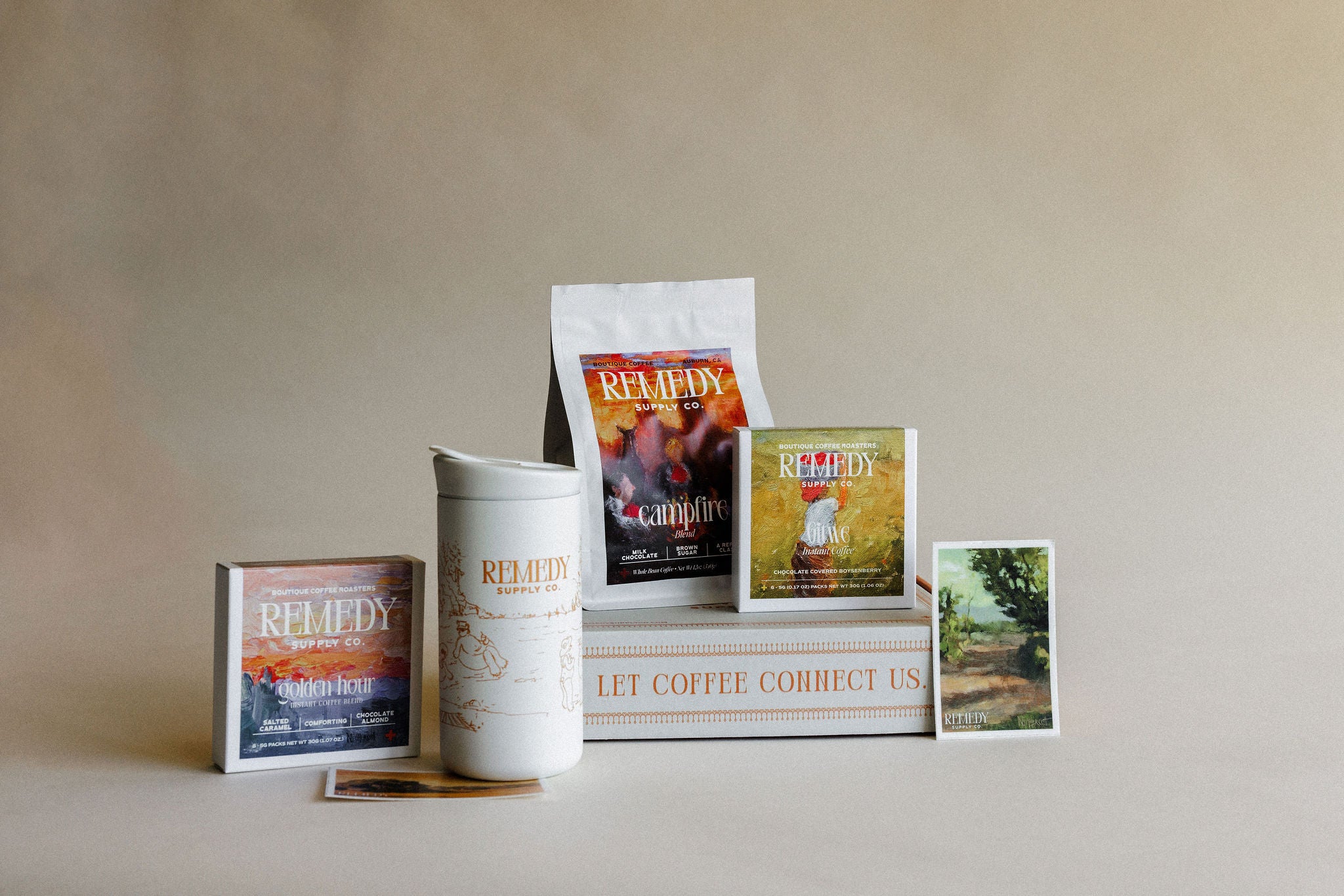 A coffee-themed gift set featuring a white ceramic tumbler, a bag of coffee, a boxed coffee gift, a small card, and a book or box with coffee-related text, arranged on a beige surface against a matching beige background.
